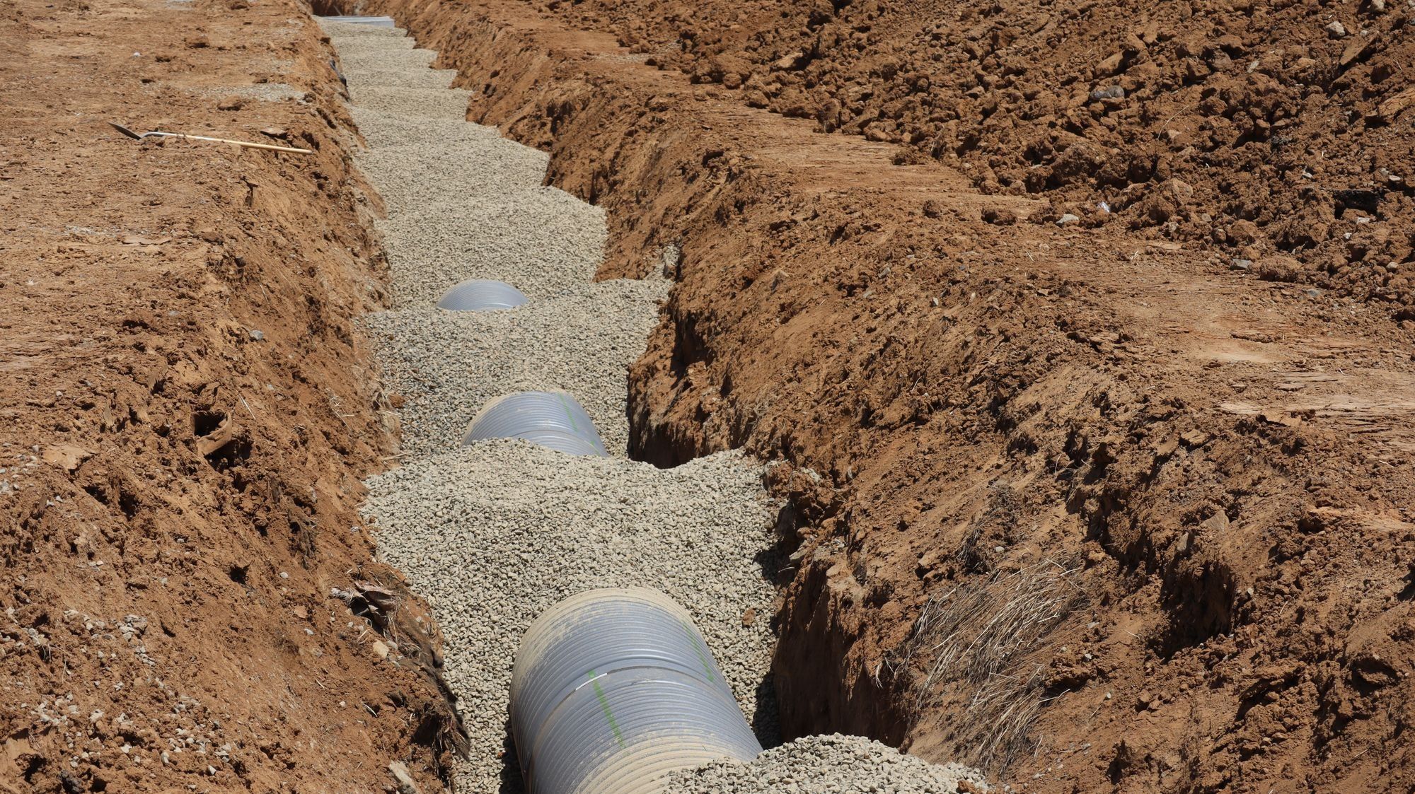 A long trench dug through clay soil, lined with gravel and part-filled with large perforated drainage pipe