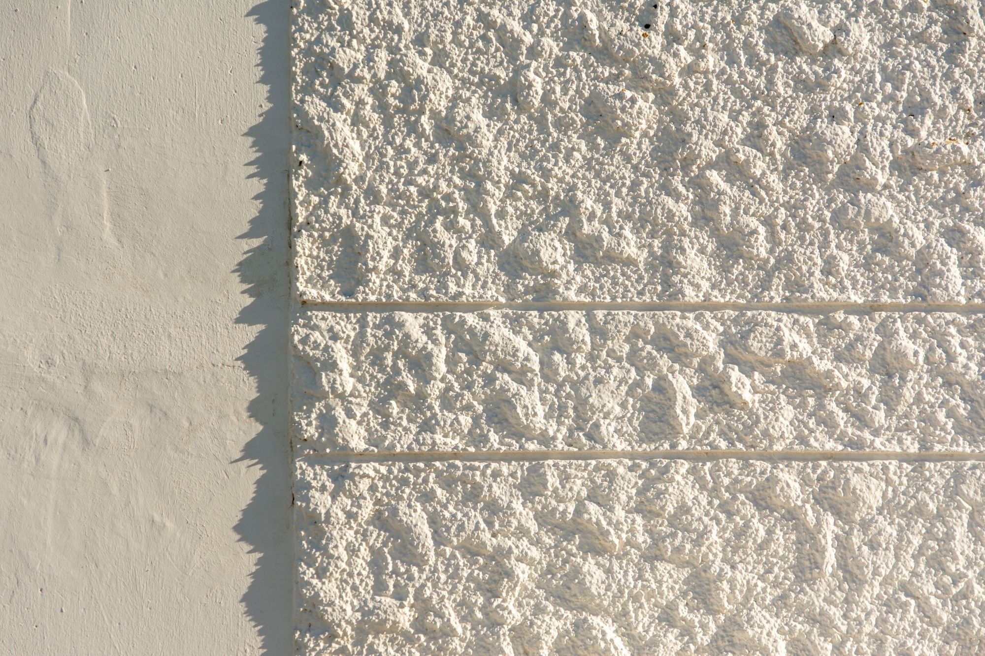 Close-up of a white painted roughcast wall with two horizontal grooves, beside a smooth painted render panel in low sunlight