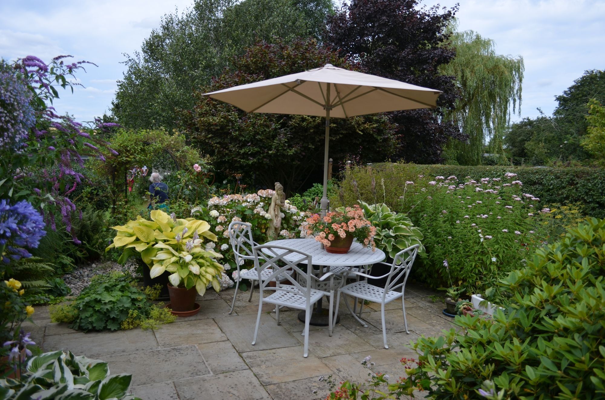 A sandstone patio with a table and parasol, surrounded by colourful cottage garden planting