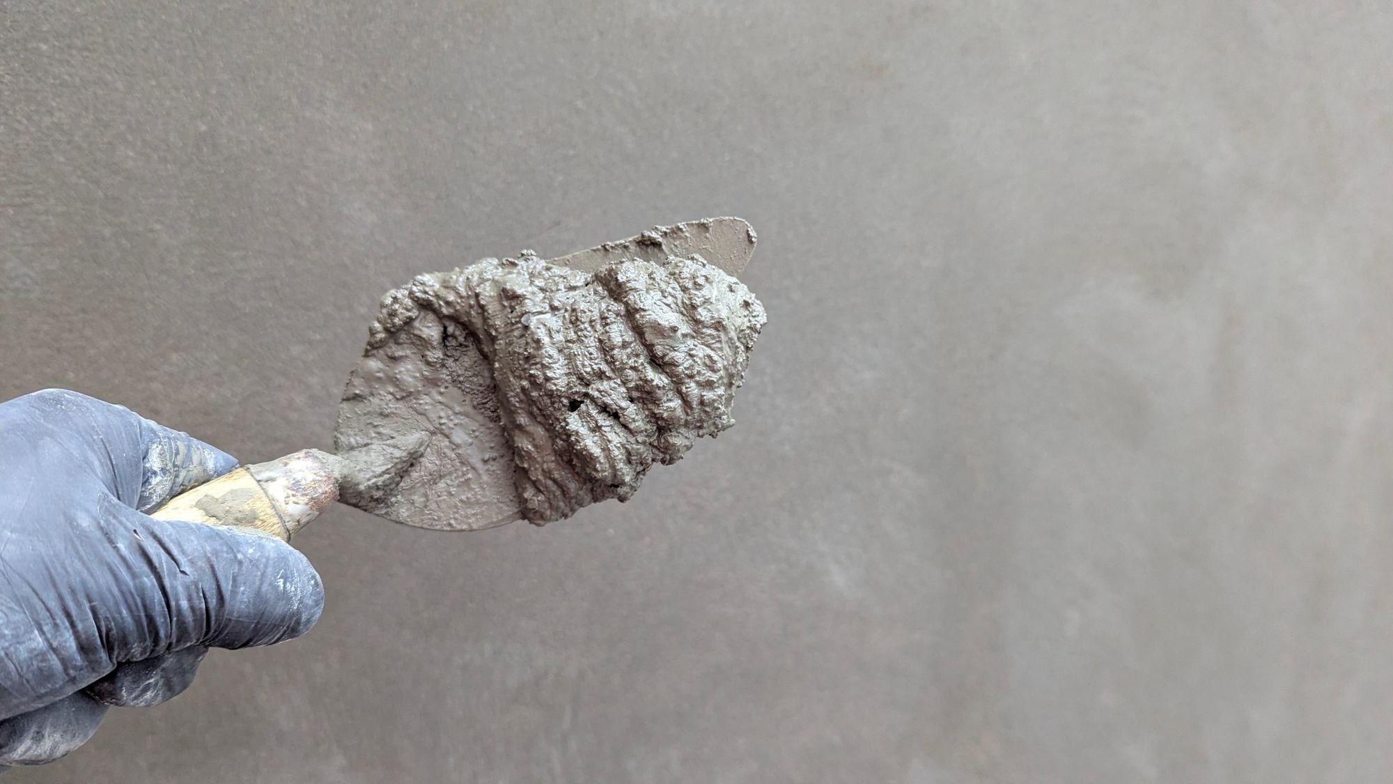 A gloved hand holding a pointing trowel loaded with wet cement mortar against a grey rendered wall